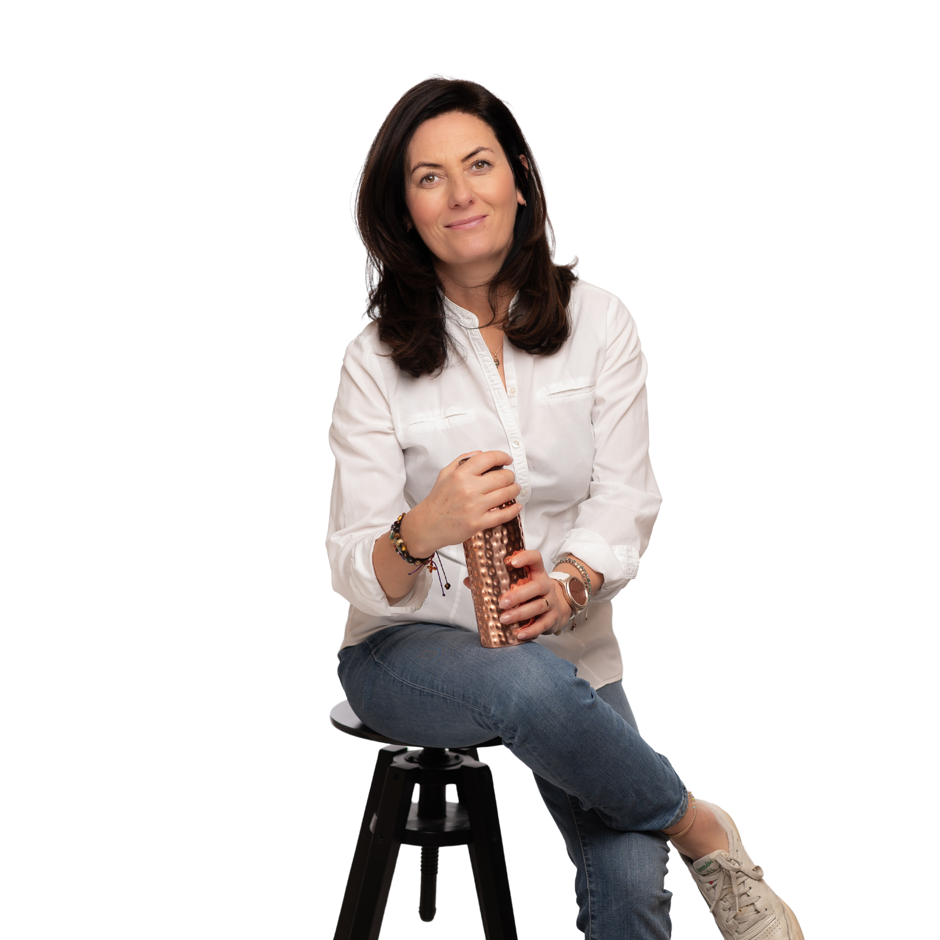 Tekst alternatywny: Uśmiechnięta kobieta w białej koszuli i niebieskich jeansach siedzi na czarnym stołku, trzymając miedzianą butelkę w dłoniach.